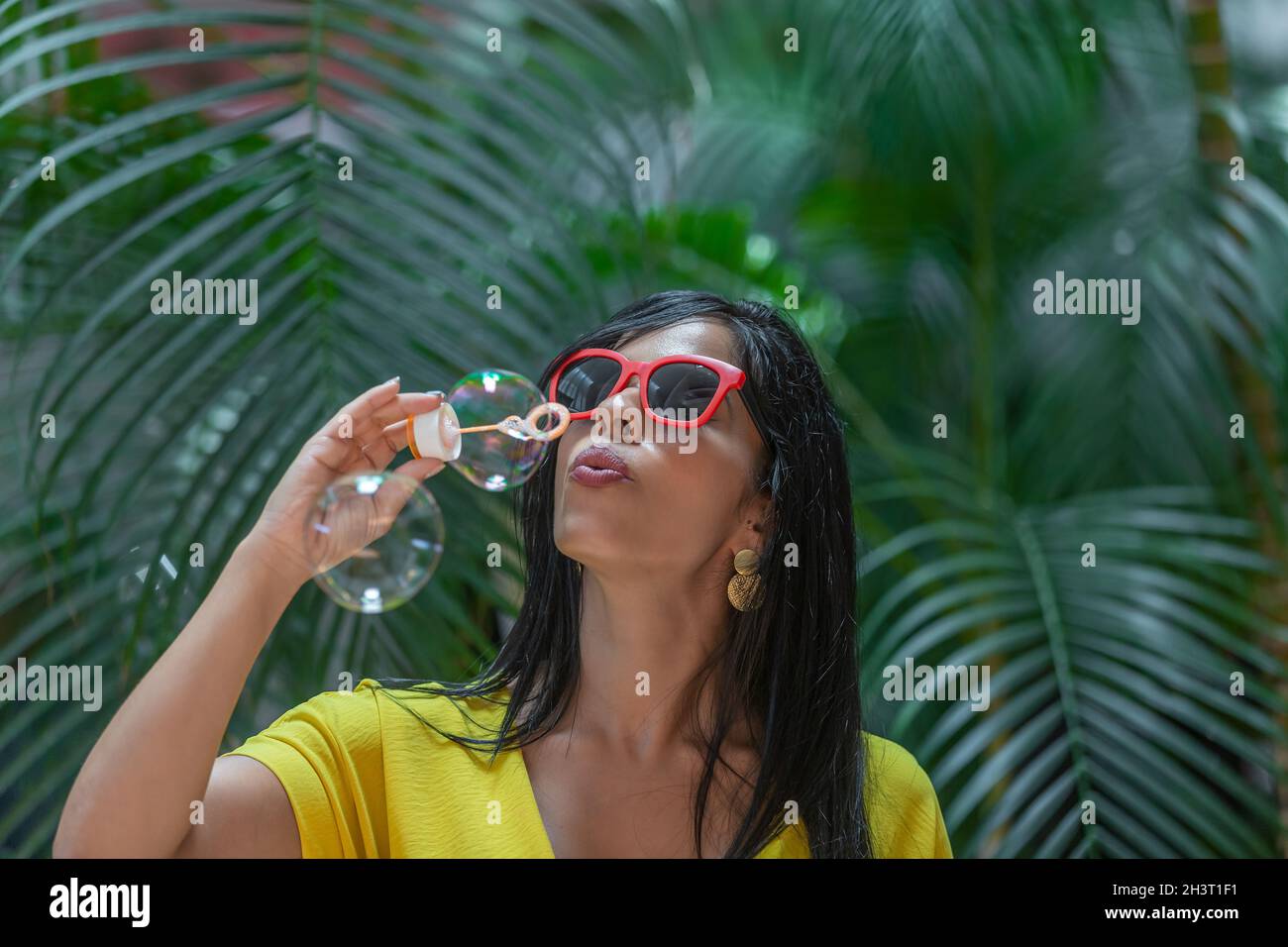 Donna latina in abito giallo e occhiali da sole con cornici rosse che soffiano bolle su sfondo di piante verdi all'aperto. Foto Stock