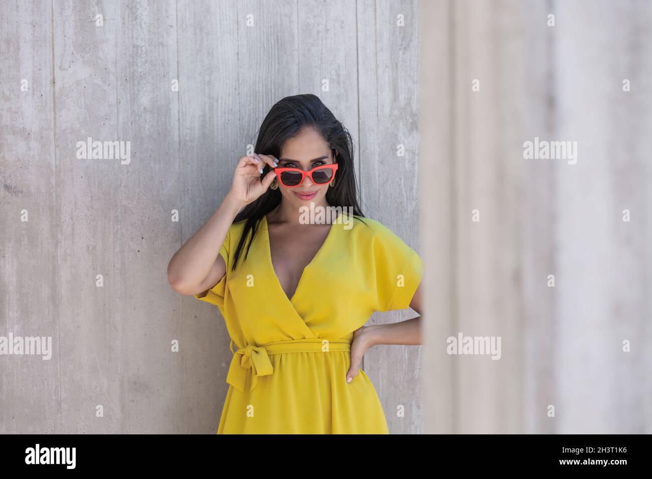 Guarda vicino a una colonna di una giovane donna latina in abito giallo indossando occhiali da sole e tenendoli con una mano e guardando la macchina fotografica sopra la parte superiore del Foto Stock