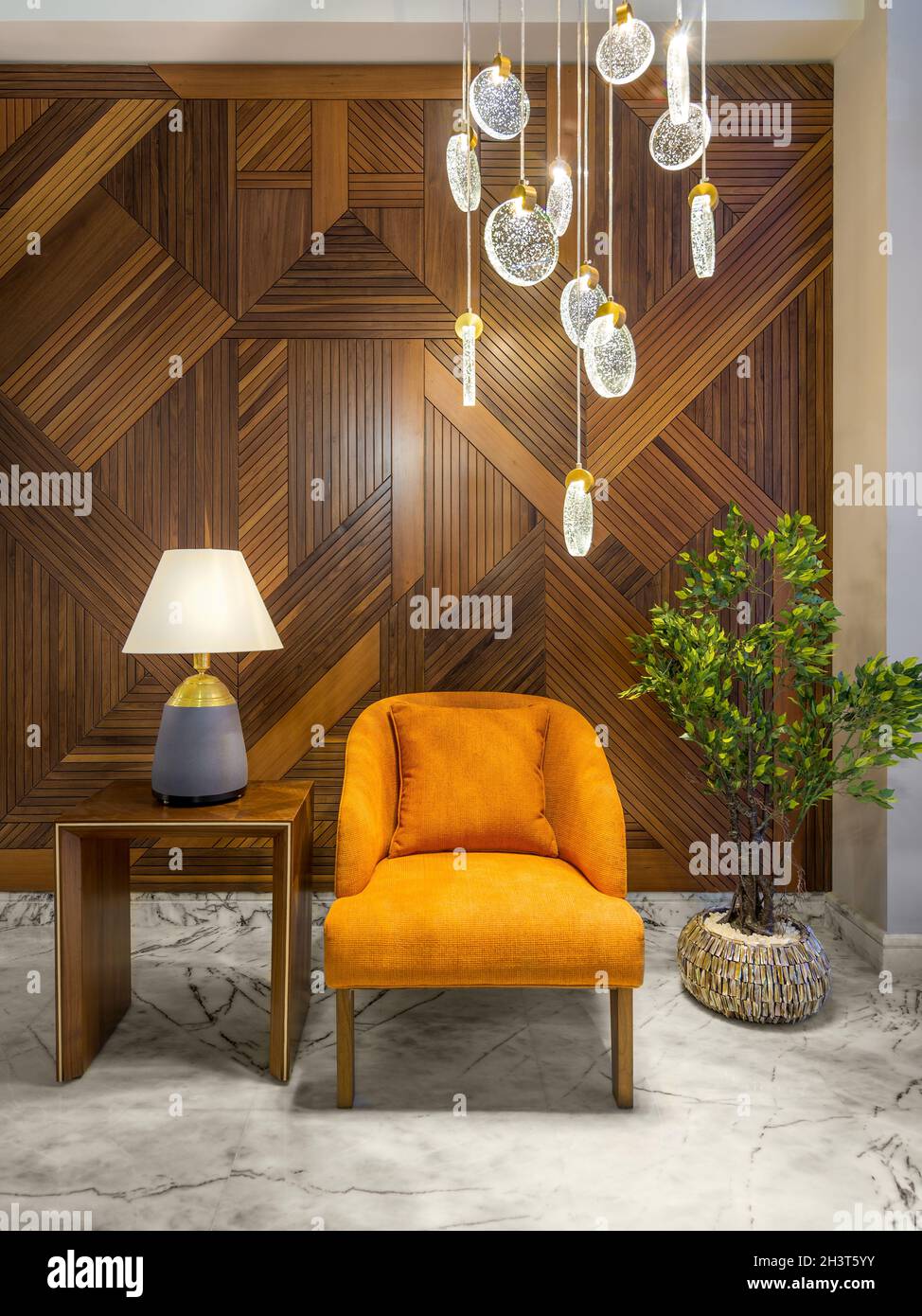 Moderna poltrona arancione, paralume lampada su un piccolo tavolo di legno, piantatrice con cespugli verdi e alto lampadario di vetro contemporaneo, in una sala con pareti rivestite di legno decorate, e pavimento di marmo bianco Foto Stock