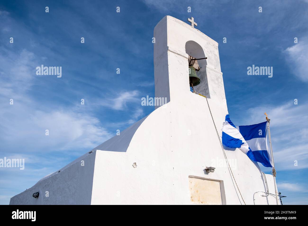 Isola di Serifos, Grecia. Piccola cappella antica, pareti bianche e campanile, bandiera greca che ondola sul muro della chiesa, cielo blu nuvoloso sfondo. Foto Stock