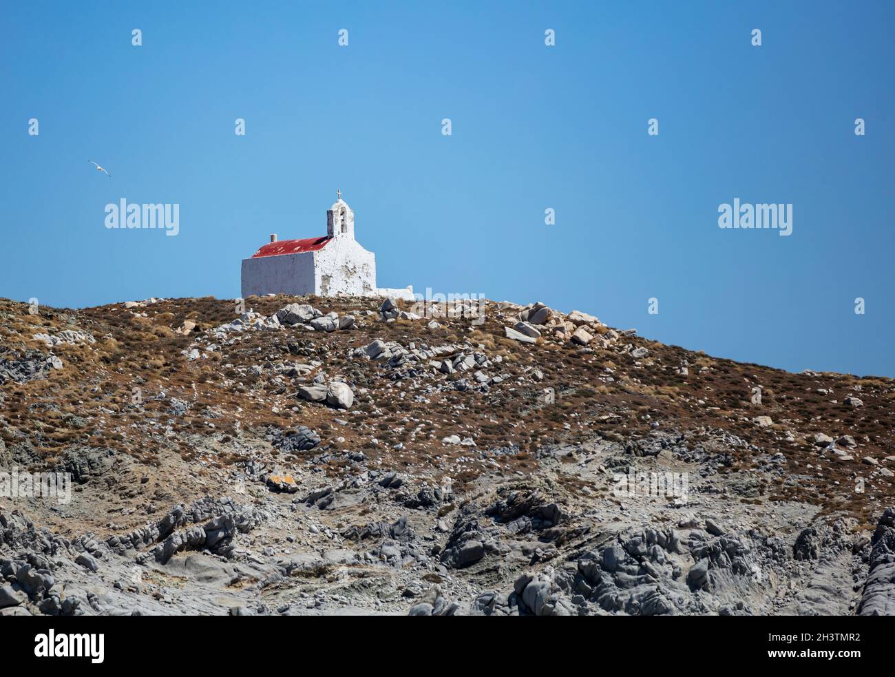 Piccola cappella antica, pareti bianche sbiadite e campanile, tetto rosso. Chiesa greca ortodossa sulla collina rocciosa, cielo blu chiaro sfondo. Isola di Mykonos, Grecia Foto Stock