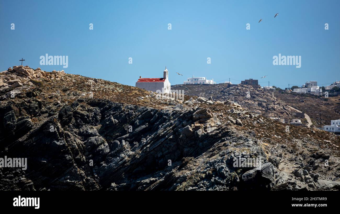 Piccola cappella antica, pareti bianche sbiadite e campanile, tetto rosso. Chiesa greca ortodossa sulla collina rocciosa, cielo blu chiaro sfondo. Isola di Mykonos, Grecia Foto Stock