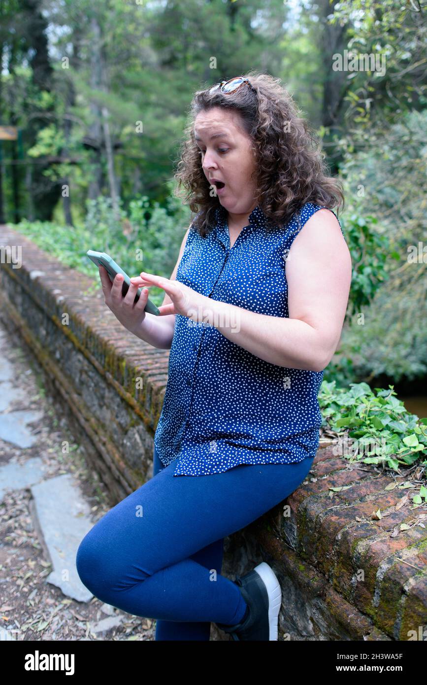 Donna caucasica, capelli tirati indietro con un paio di occhiali da sole e un volto sorpreso guardando lo smartphone in una foresta, mentre poggiato contro di me Foto Stock