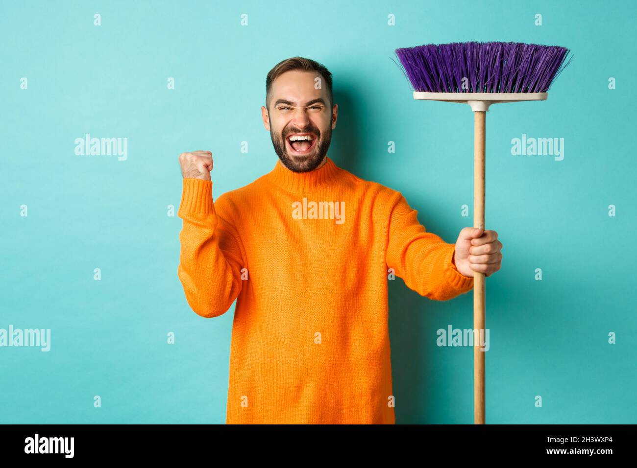 Immagine di un uomo incoraggiato e motivato che si prepara per la pulizia, la scopa in mano e il gesto di fare il pugno, in piedi sopra turq Foto Stock