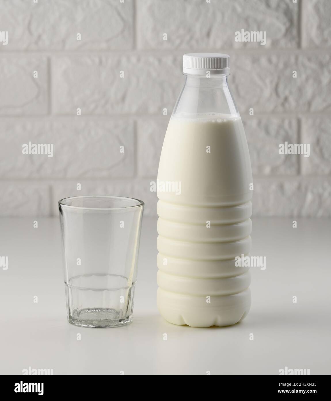 Bottiglia di latte in plastica piena e tazza di vetro vuota su un tavolo bianco, sfondo in mattoni Foto Stock