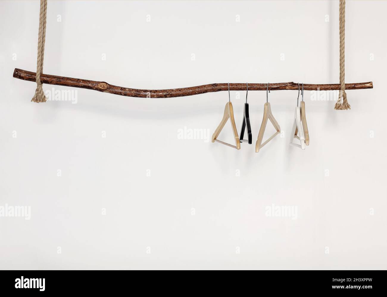 Cinque appendiabiti vuoti appesi su un bastone in legno rustico su una corda spessa, isolato su una parete bianca Foto Stock