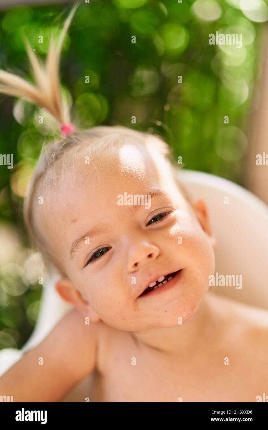 Una bambina sorridente con coda di ponile si siede su una sedia alta. Verticale Foto Stock