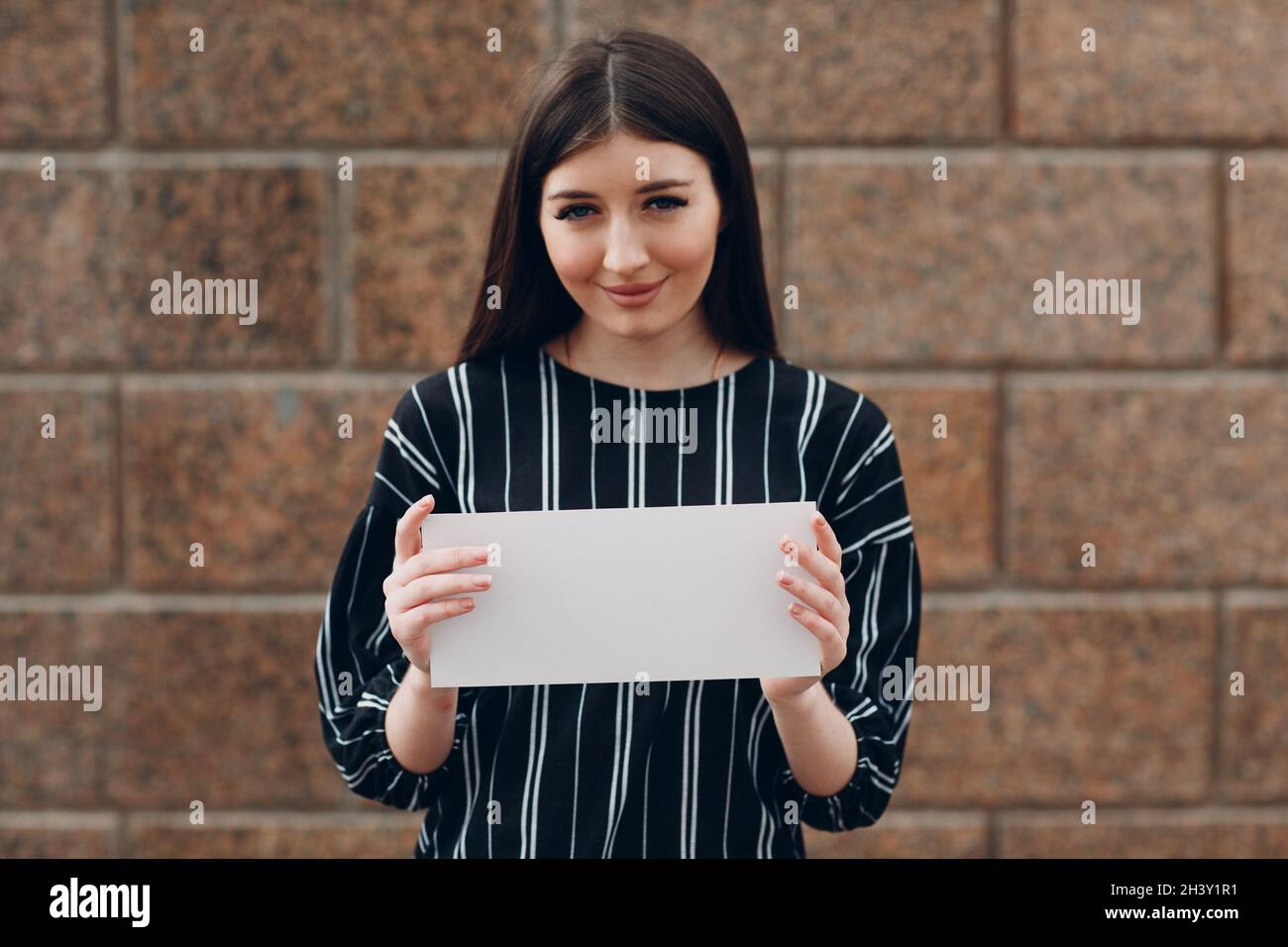 Giovane donna con sorriso tiene carta bianca in mano pietra sfondo. Ragazza con foglio bianco modello con spazio vuoto. Foto Stock