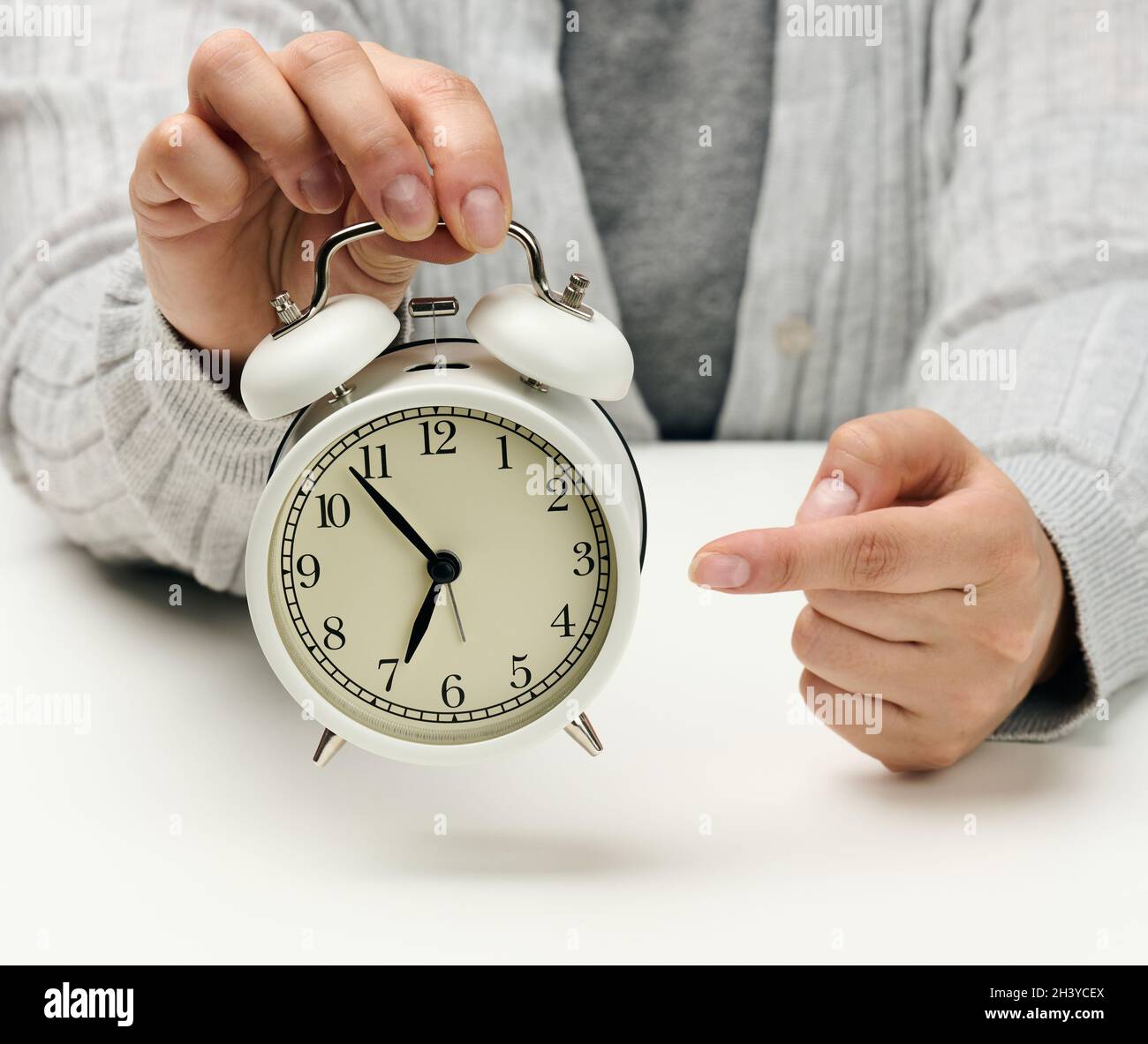 La mano femminile tiene una sveglia in metallo bianco su un tavolo bianco, tempo cinque minuti a sette al mattino Foto Stock