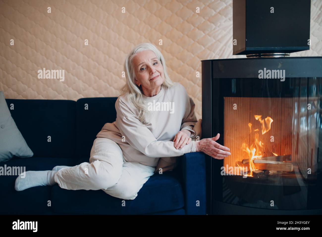 Donna anziana con capelli grigi seduta sul divano in soggiorno con camino Foto Stock