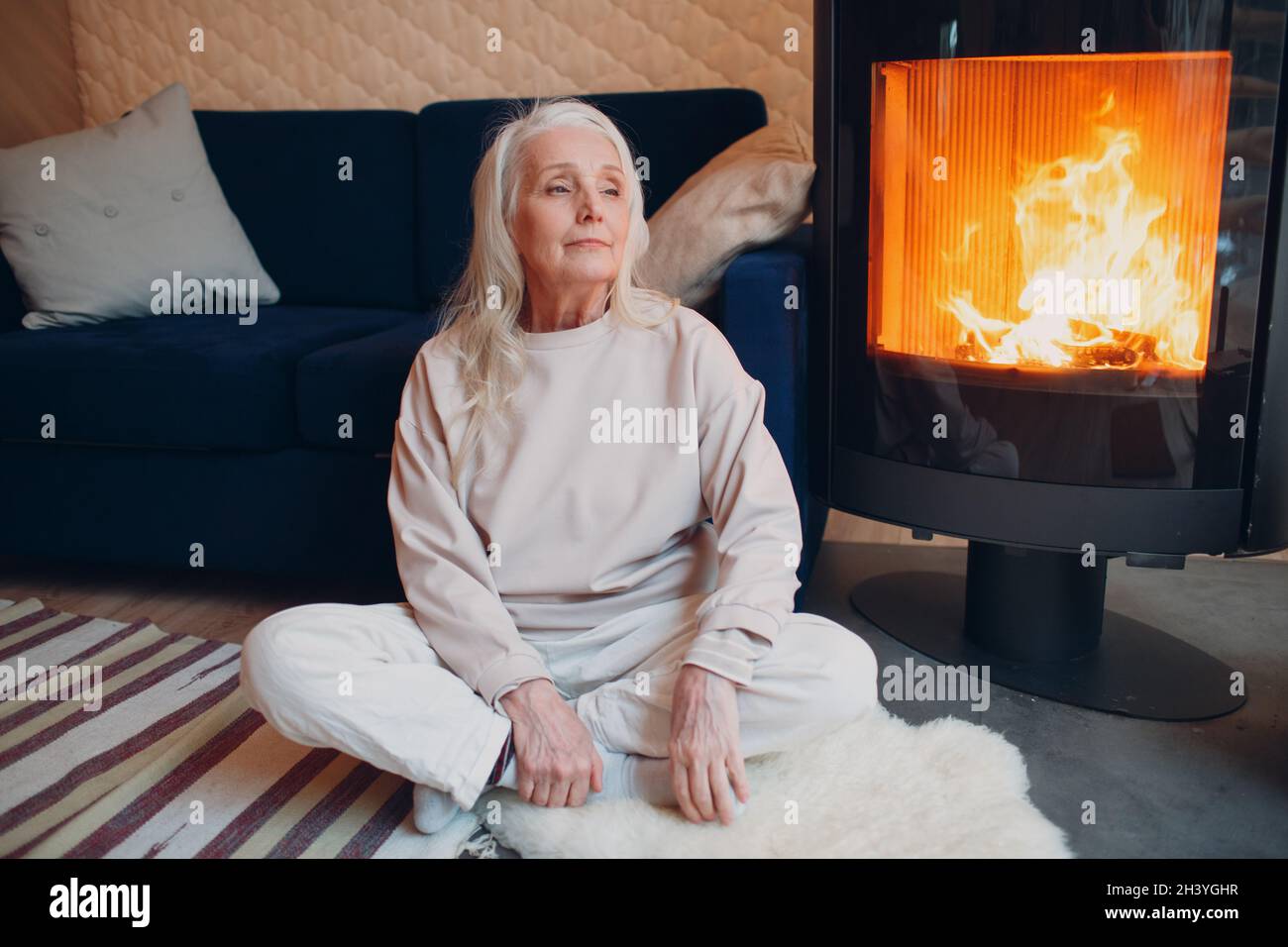 Donna con capelli grigi mature seduta su divano in soggiorno con camino Foto Stock