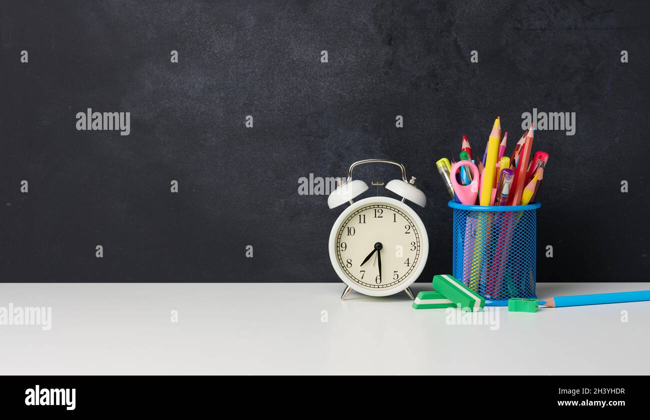 Sveglia rotonda bianca e un vetro di metallo con penne, matite e pennarelli sullo sfondo di un bordo nero vuoto di gesso Foto Stock
