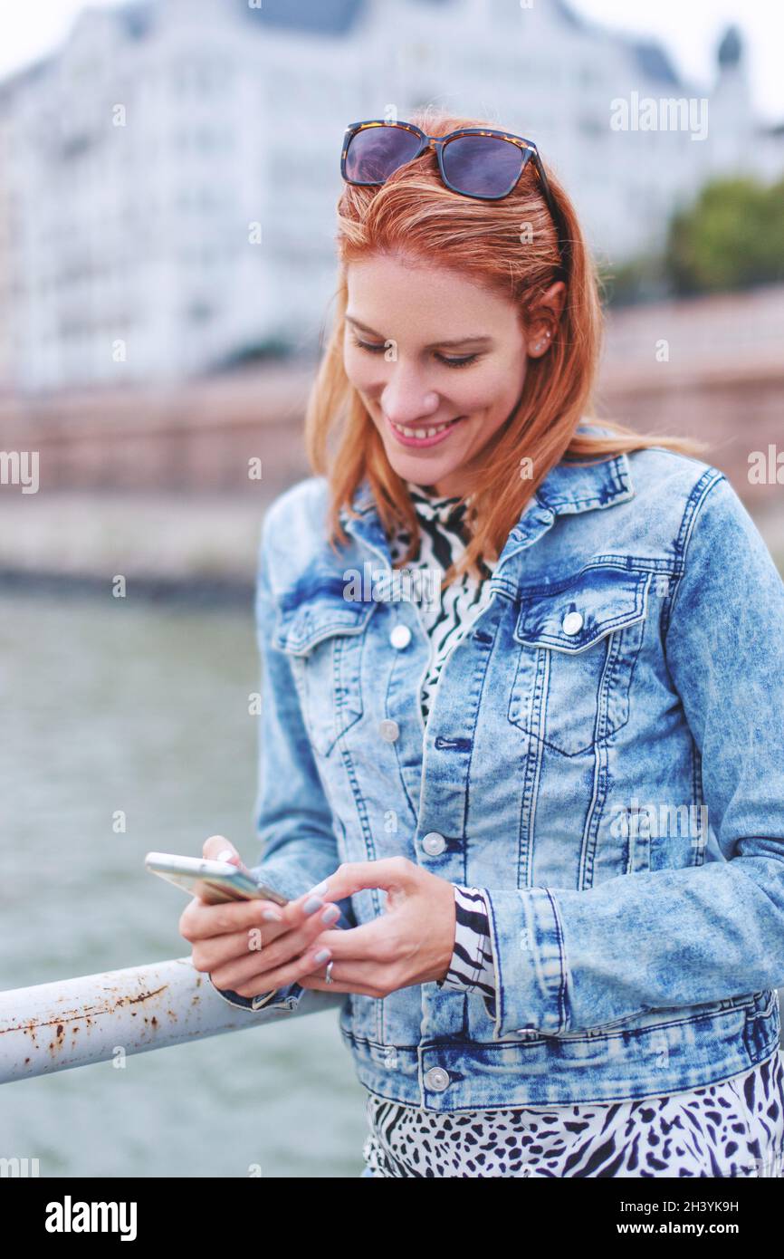 Felice giovane rossa caucasica donna leggere messaggio sullo smartphone in riva al fiume Foto Stock