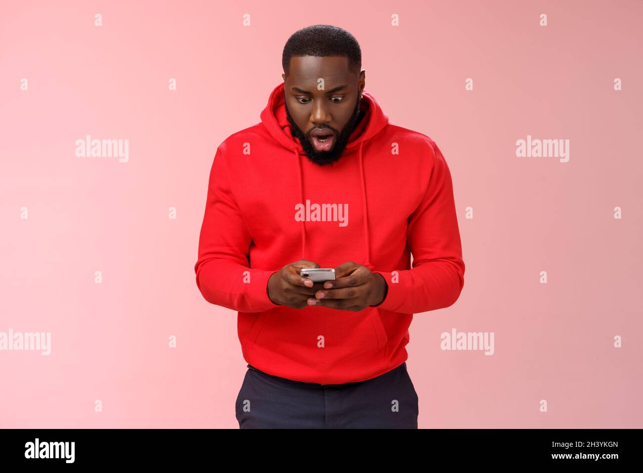 Divertito messo a fuoco impressionato afroamericano bearded ragazzo in rosso hoodie goccia mascella occhi allargati sguardo stupito smartphone display vincendo h Foto Stock