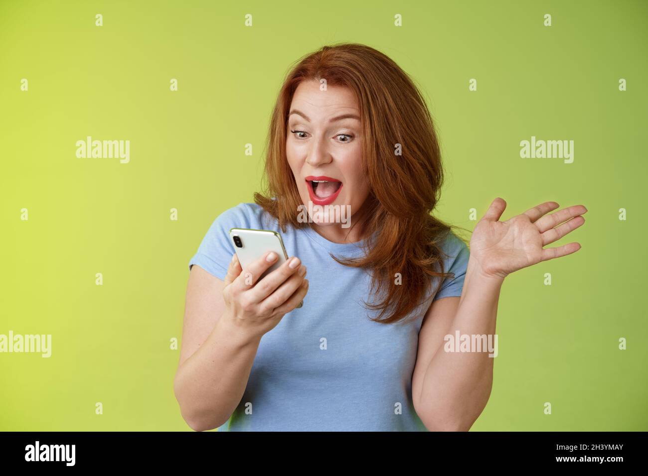 Sorpreso felice di mezza età piuttosto redhead donna guardare smartphone sollevare mano celebrativa eccitazione bocca aperta reazione impressionato Foto Stock