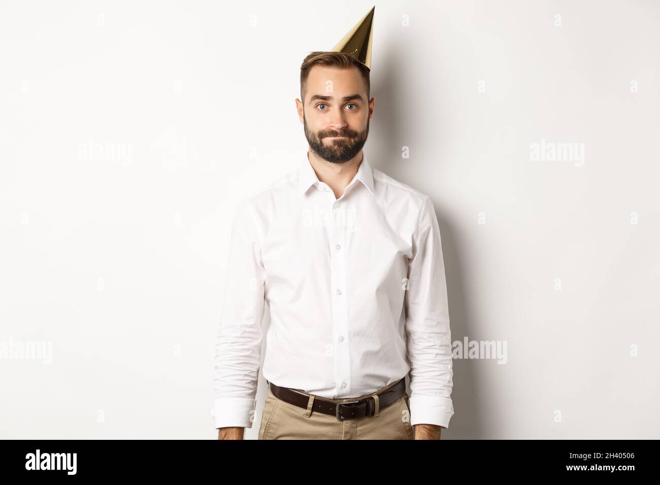 Festeggiamenti e feste. Ragazzo cupo in cappello di compleanno in piedi scomodo su sfondo bianco, sentendosi indivertito Foto Stock