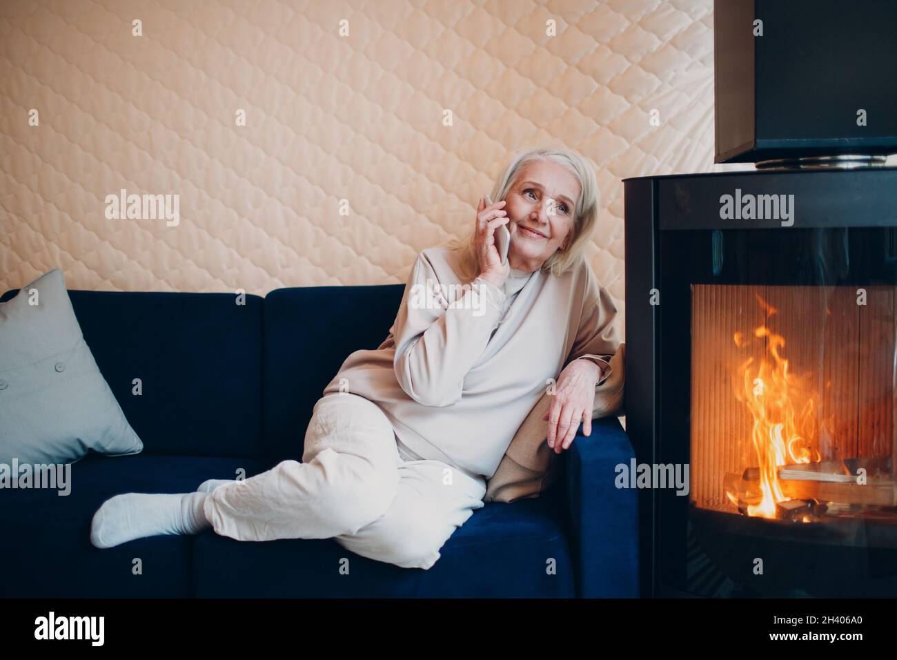 Donna anziana con capelli grigi seduta sul divano e smartphone parlante in soggiorno con camino Foto Stock