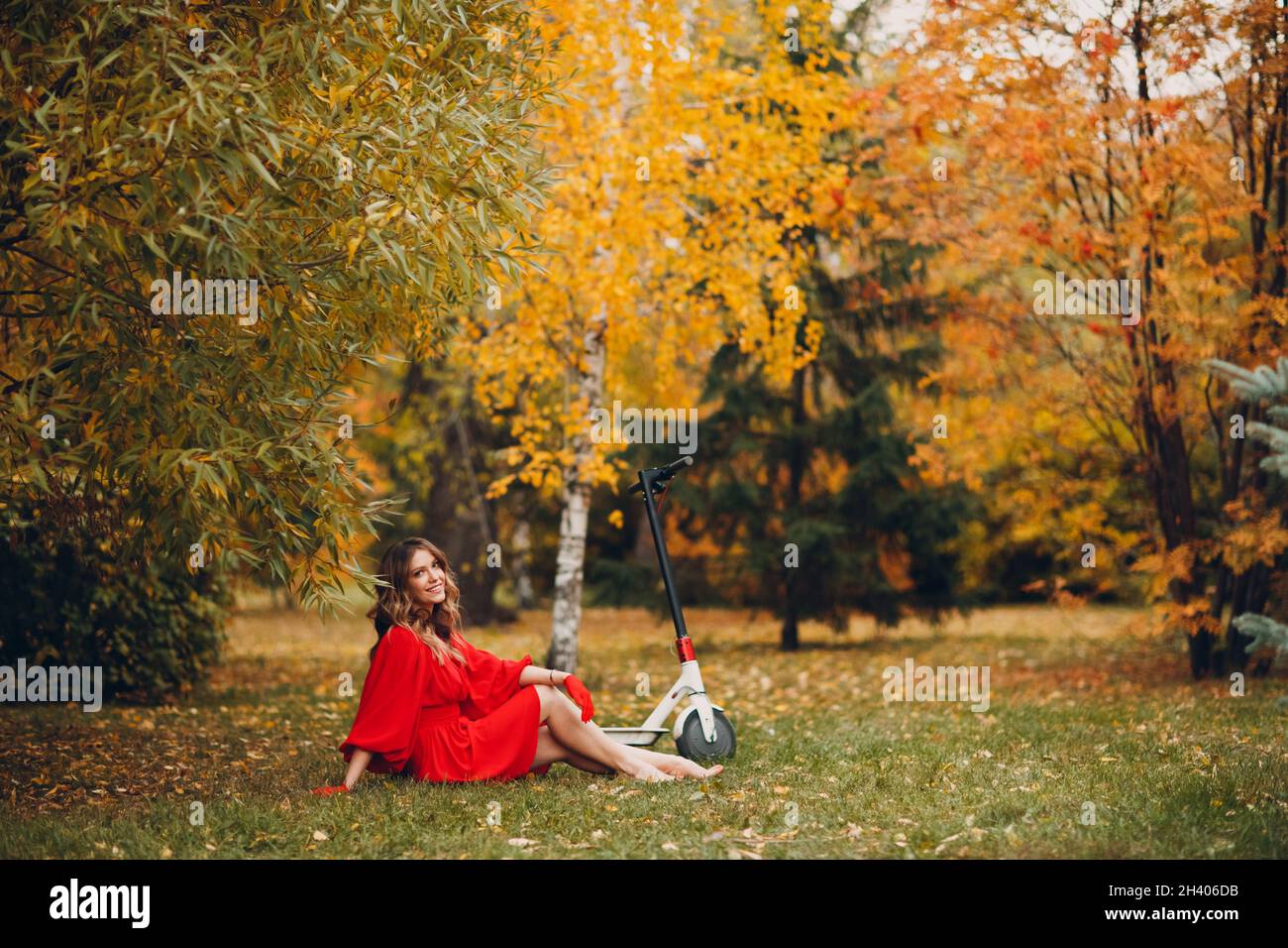 Giovane donna seduta con scooter elettrico in abito rosso a. il parco della città d'autunno Foto Stock