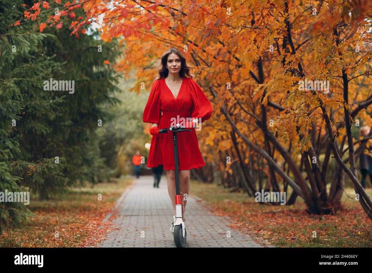 Giovane donna con scooter elettrico in abito rosso al parco cittadino d'autunno Foto Stock