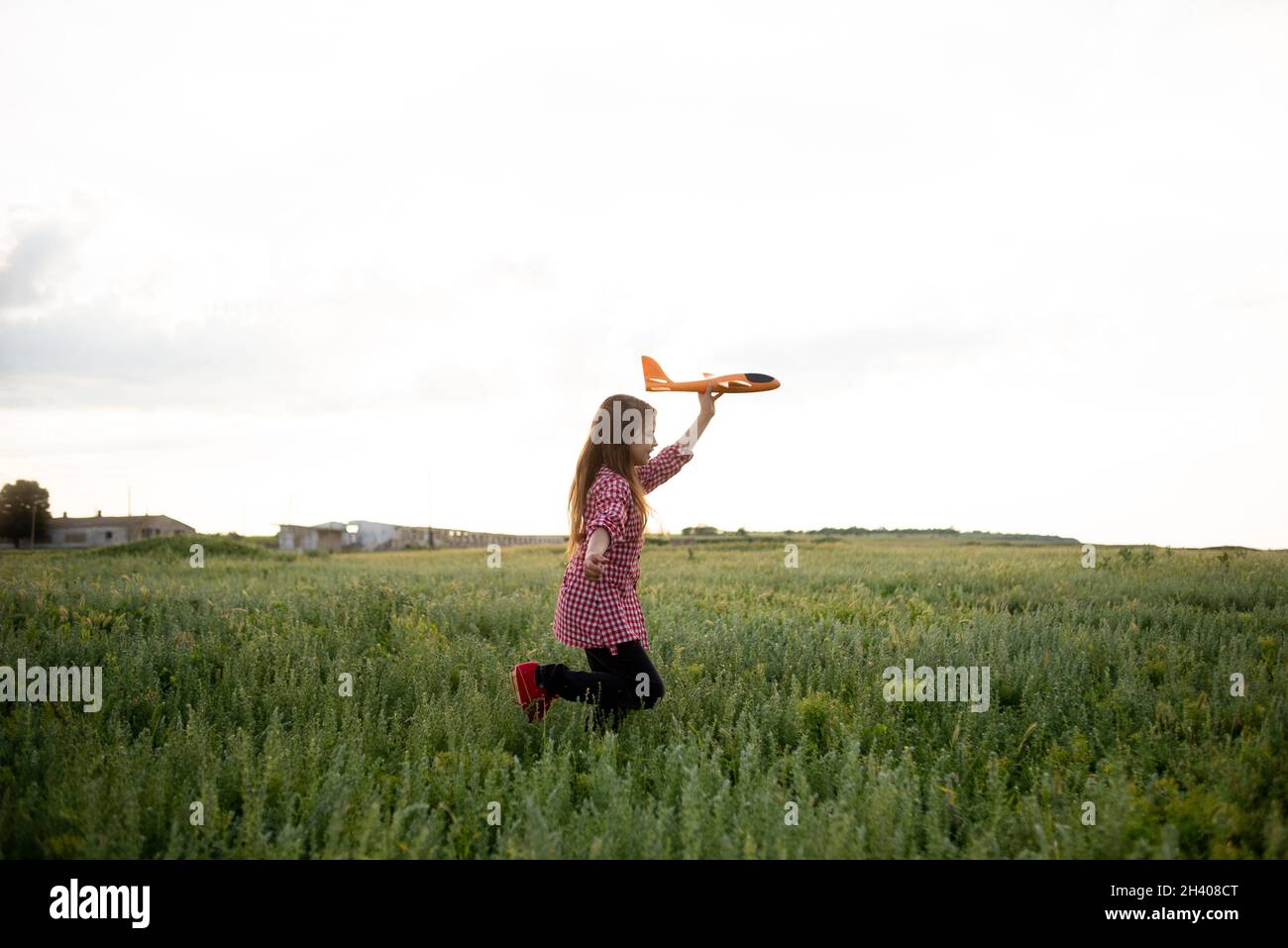 La ragazza caucasica felice che corre attraverso il campo lancia un aeroplano giocattolo nel cielo con la mano in su. Bambino gioioso cammina da solo, corre sull'erba Foto Stock