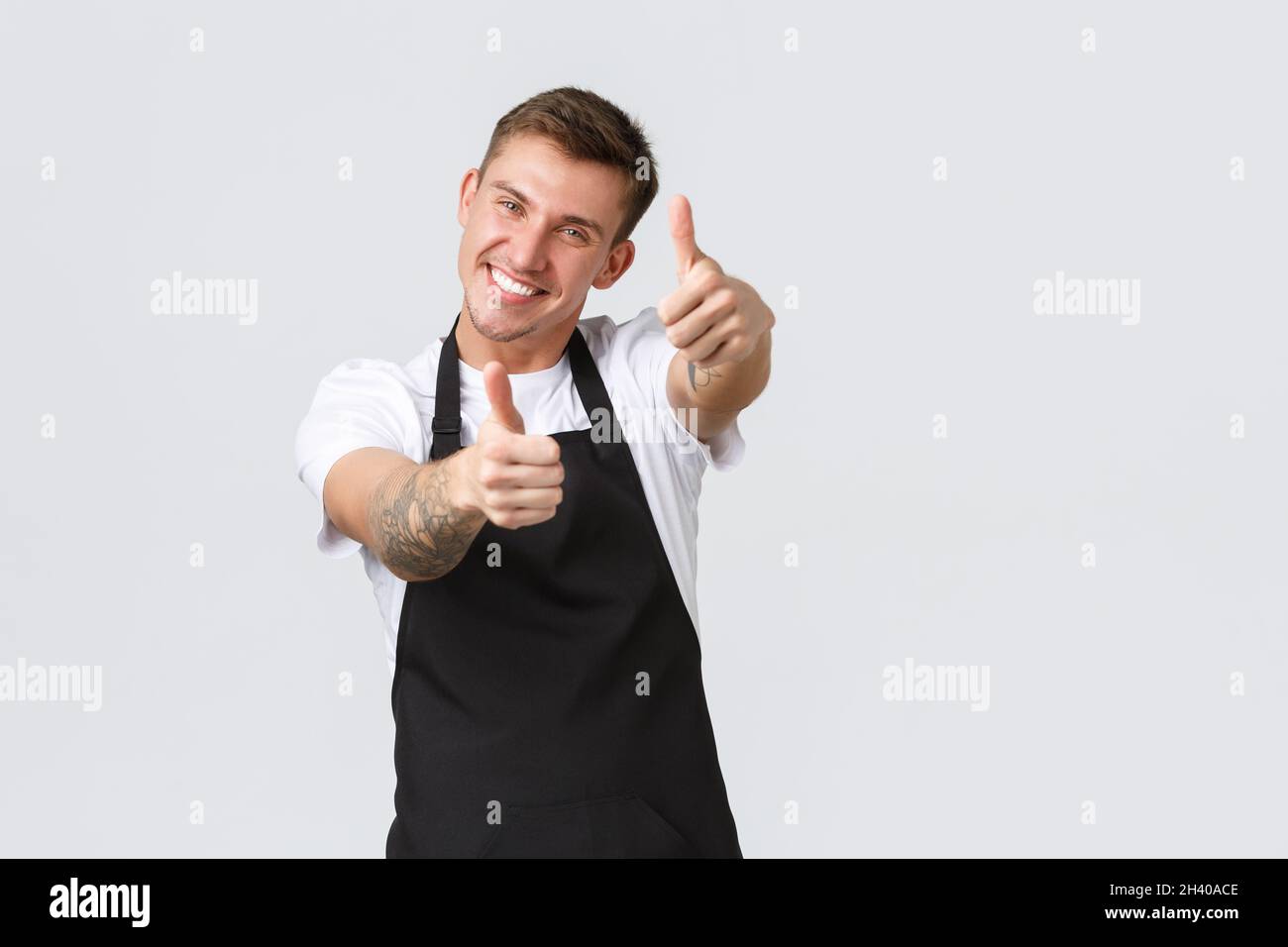 Dipendenti, negozi di alimentari e caffè concetto. Un barista allegro e bello al bar, un dipendente del negozio in grembiule nero che mostra il tum Foto Stock