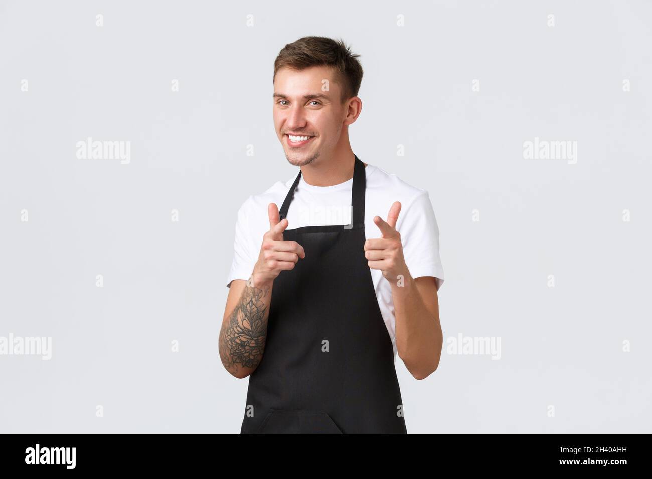 Dipendenti, negozi di alimentari e caffè concetto. Carino cameriere biondo, barista in grembiule nero che sorride ampiamente e poin Foto Stock