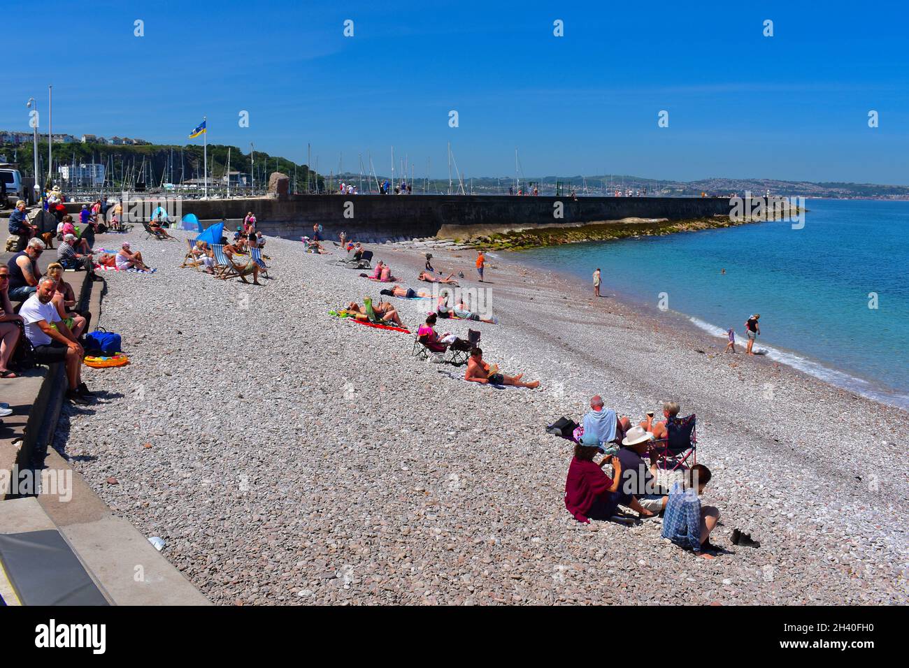 La gente si diverta a sedersi e giocare sulla spiaggia ciottolosa di Breakwater all'esterno di Brixham Marina. Foto Stock