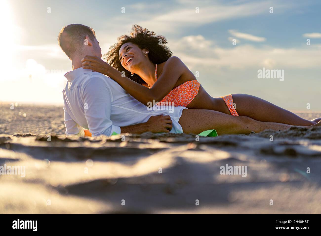 Una giovane coppia romantica multirazziale si carezza teneramente l'un l'altro sdraiato sulla spiaggia al tramonto - coppia multirazziale romantica sulla spiaggia - Amore e tra Foto Stock