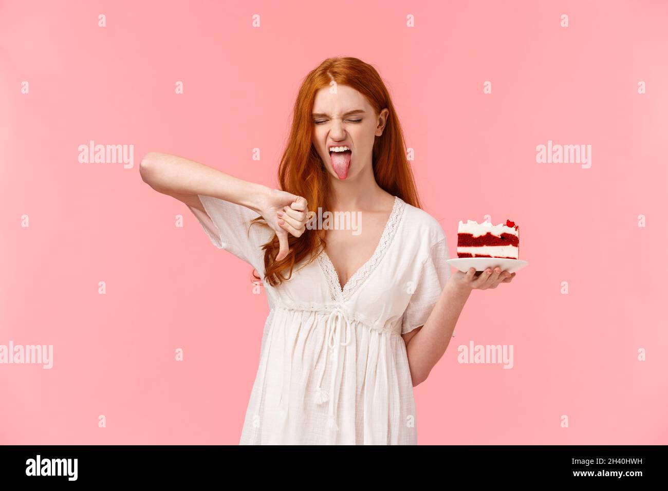 Ignorante, sottaceto e snobbish redhead compleanno ragazza odio b-giorno torta, tenendo piatto con dessert, mostrando lingua grimacing e mak Foto Stock