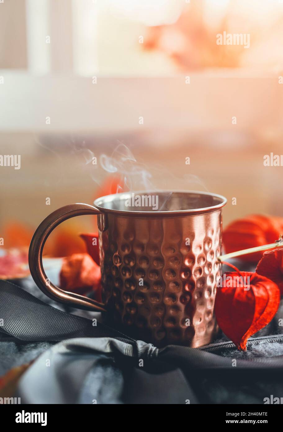 Tazza di rame con tè fumante su lana grigia con foglie d'autunno d'arancio e phisalis sullo sfondo della finestra con luce naturale. Accogliente periodo autunnale a casa Foto Stock