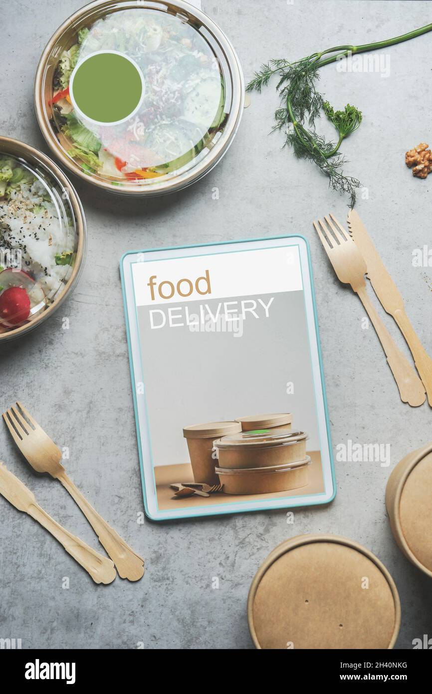 Tablet PC con display per la consegna degli alimenti su tavolo da cucina in cemento grigio chiaro con vaschette e posate in plastica. Ordine moderno cibo sano online. T Foto Stock