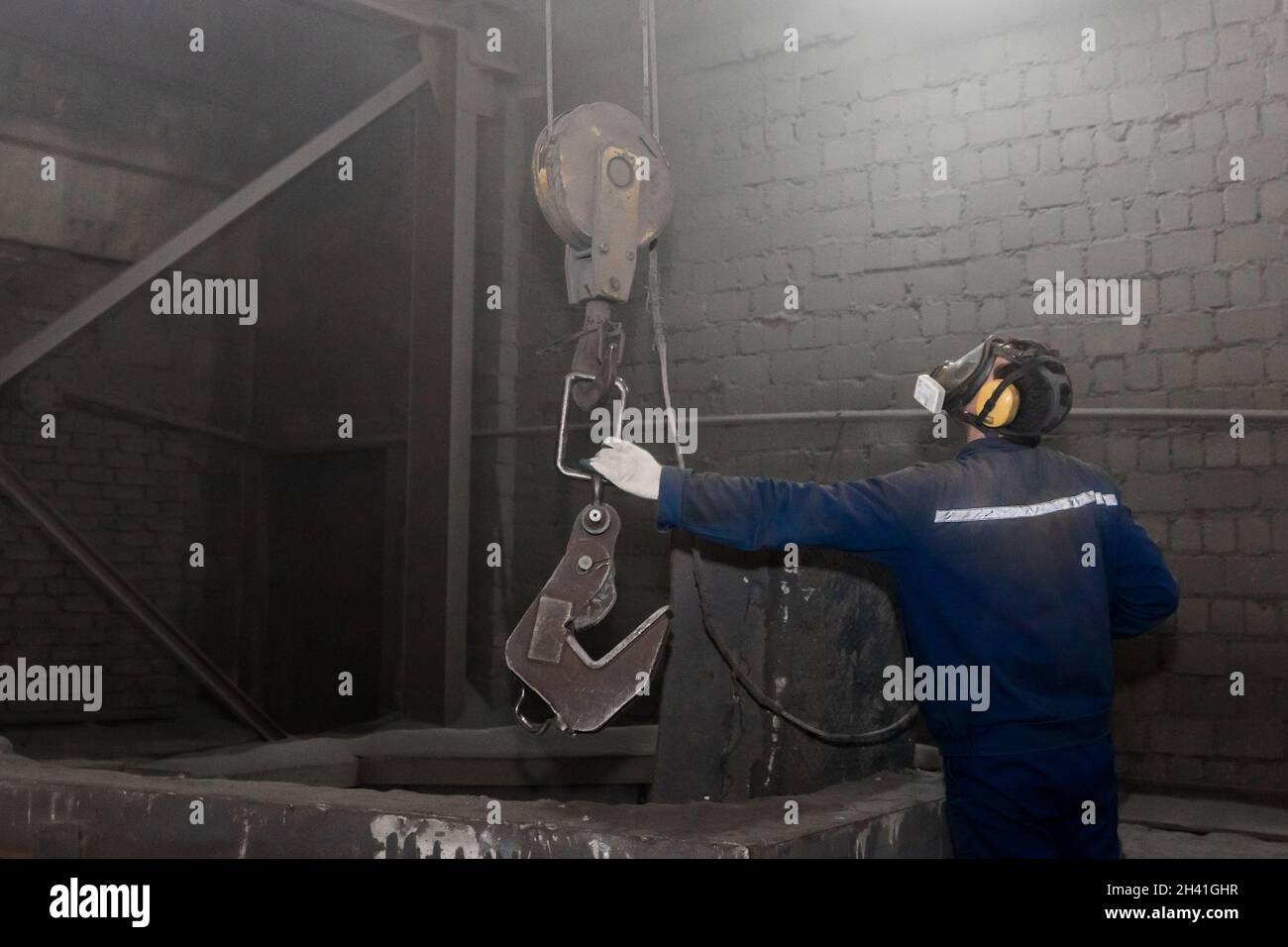 Un uomo in una tuta e respiratore che lavora prende un gancio con la mano un meccanismo di sollevamento industriale in officina di fabbrica. Foto Stock