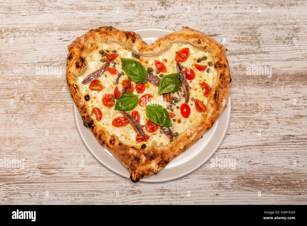 Pizza a forma di cuore con acciughe in scatola, foglie di basilico, pomodori ciliegini e mozzarella Foto Stock