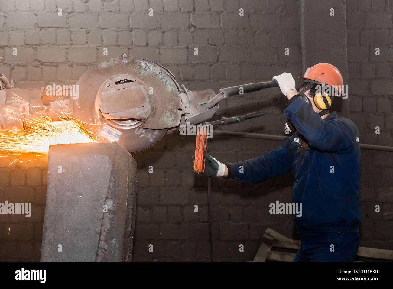 Un operatore maschile in un casco protettivo, respiratore, tute gestisce attrezzature di molatura pesanti per tubi in ghisa con scintille volanti nel wo Foto Stock