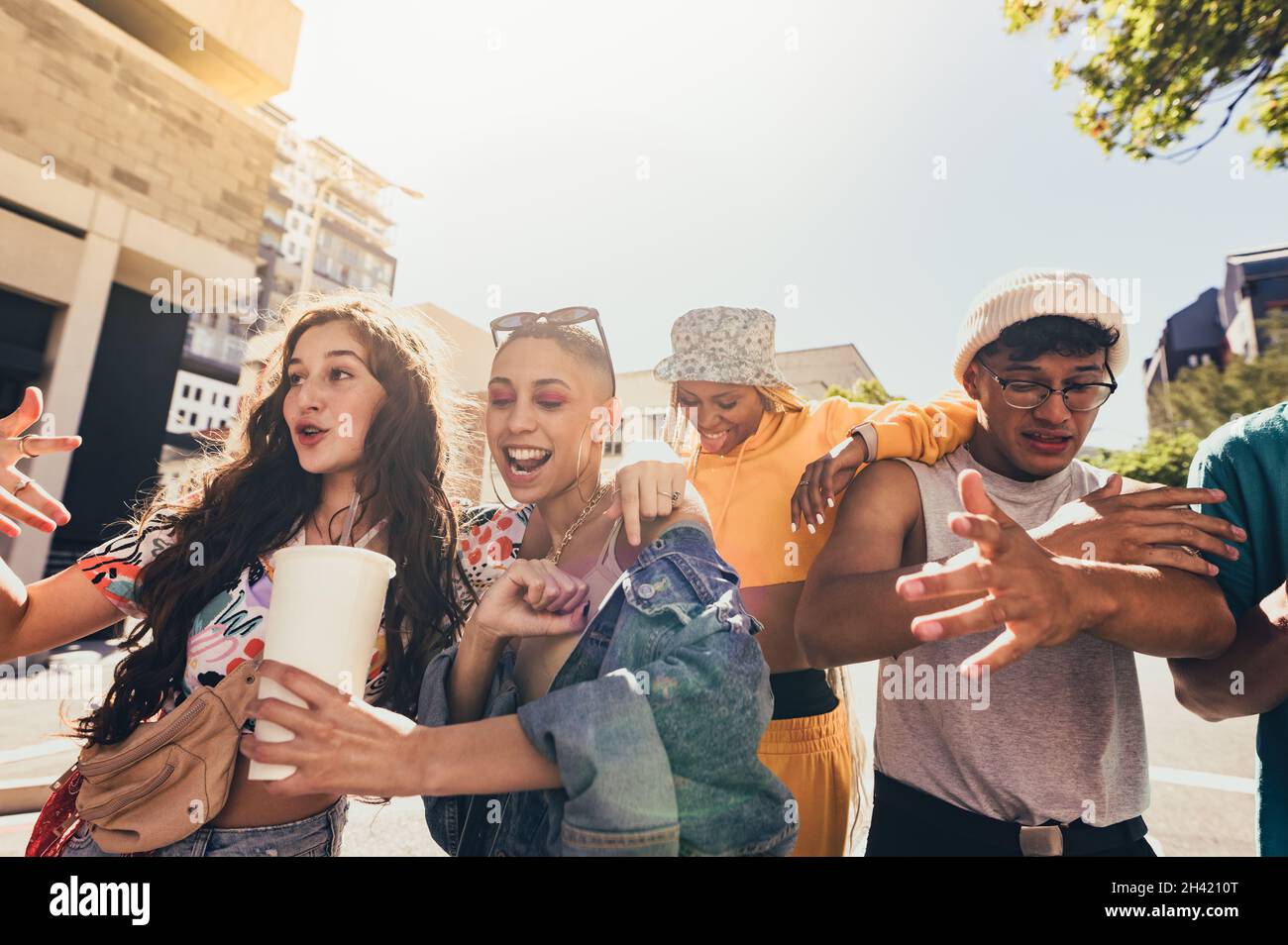 Balli in città. Gruppo di amici di generazione z cantano e ballano mentre camminano insieme nel sole estivo. Amici giovani felici che si divertono mentre Foto Stock