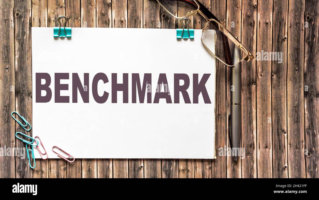 Benchmark, testo su blocco note bianco. Occhiali e fermagli di carta su un tavolo di legno. Foto Stock