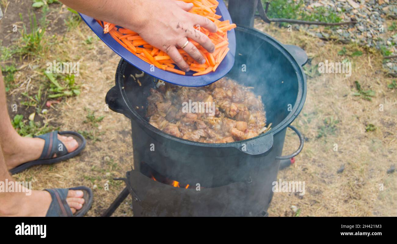 L'uomo caucasico cuoce la carne con le carote in una padella di wok su un fuoco aperto, estate, all'aperto. Foto Stock