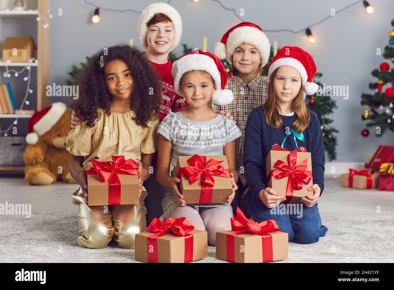 Gruppo di bambini felici e diversi che tengono regali di Natale seduti sul pavimento a casa Foto Stock