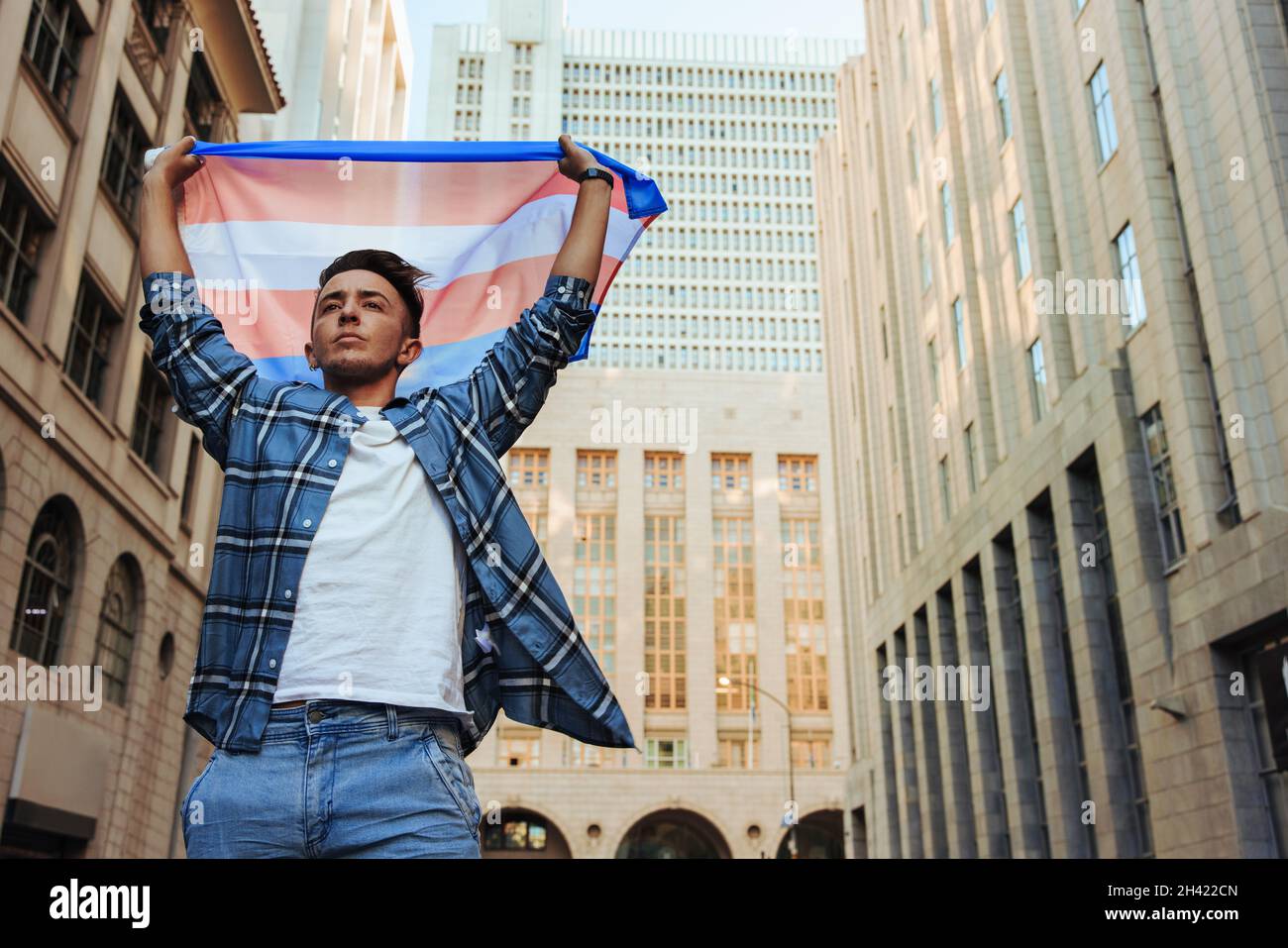 Uomo non conforme che alza la bandiera transgender all'aperto. Giovane uomo transgender fiducioso che celebra l'orgoglio gay nella città. Sesso giovane non conforme Foto Stock