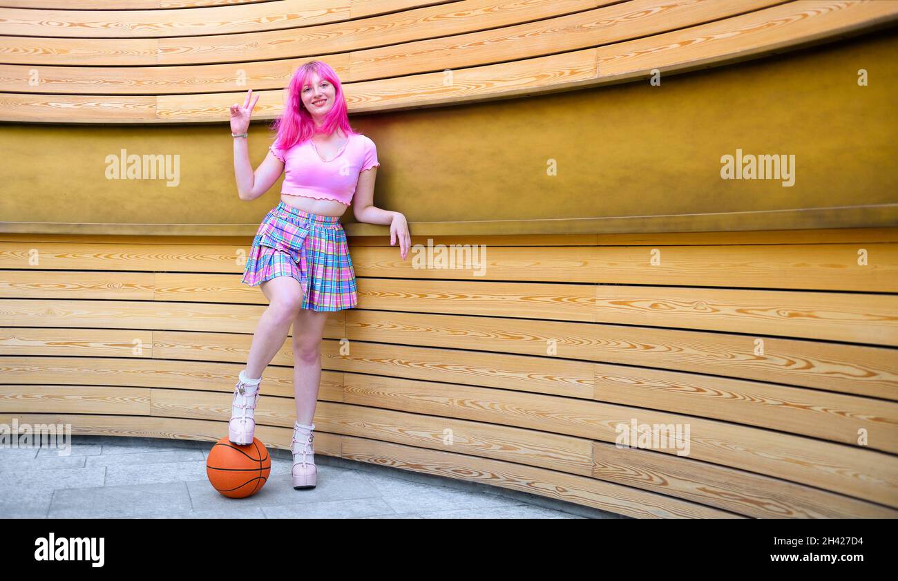 Una bella ragazza con capelli rosa in posa di fronte al muro - Donna millennial in piedi in urbanity area pubblicitaria con il piede poggiato sul bask Foto Stock