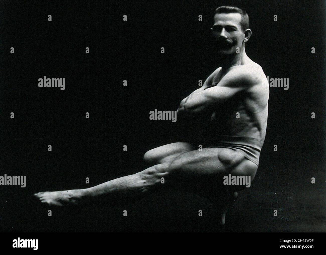 Un bodybuilder maschile che indossa tronchi da bagno, che colpisce una posa 'Cossack dance', in un ambiente studio. Fotografia di Hana Studios, ca.1899-1908. Foto Stock