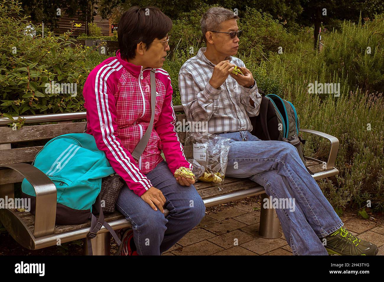 Due amici maschi seduti su una panca in un parco che mangiano sandwich. Foto Stock