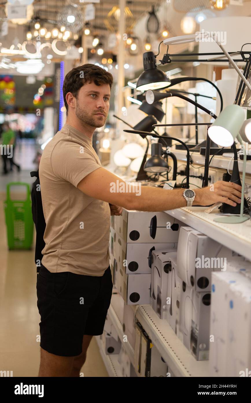 Vista laterale di un uomo casual bello guardando di lampade da tavolo su scaffale e selezionarne uno mentre si acquista nel reparto home Goods Foto Stock