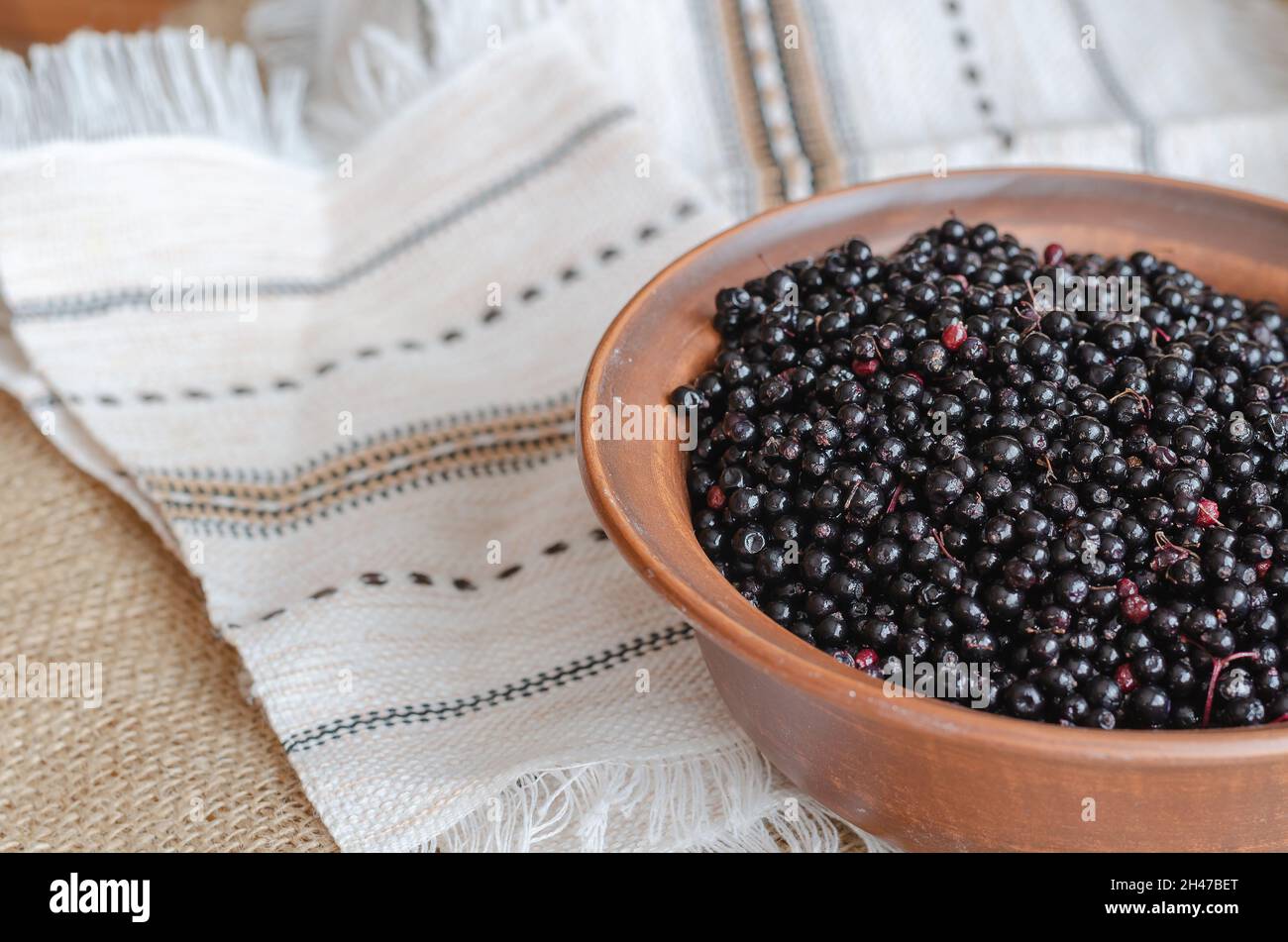 Sambuchi neri maturi in una ciotola marrone. Ciotola di argilla sopra una tovaglia etnica di lino. Sambucus nigra. Messa a fuoco selettiva. Foto Stock