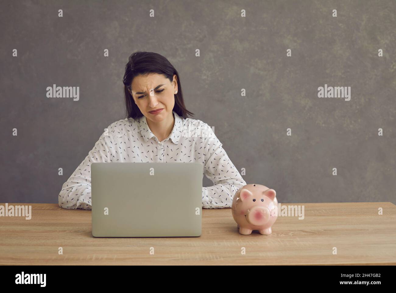 Donna triste con la banca piggy guardando lo schermo del laptop e pensando ai problemi di soldi Foto Stock