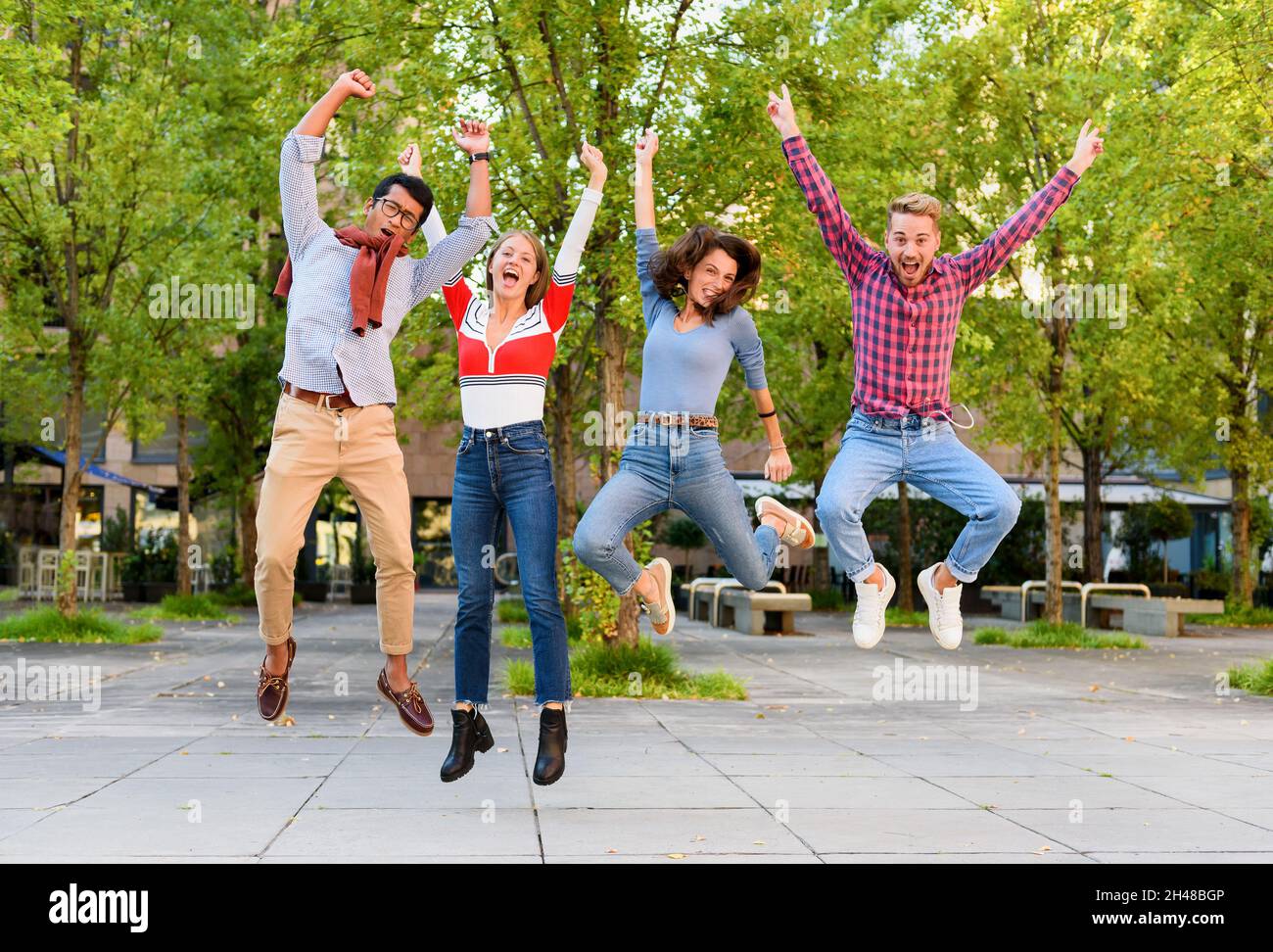 Gruppo di giovani amici esuberanti e diversi che allietano e saltano in aria con le braccia sollevate in festa all'aperto in un parco Foto Stock