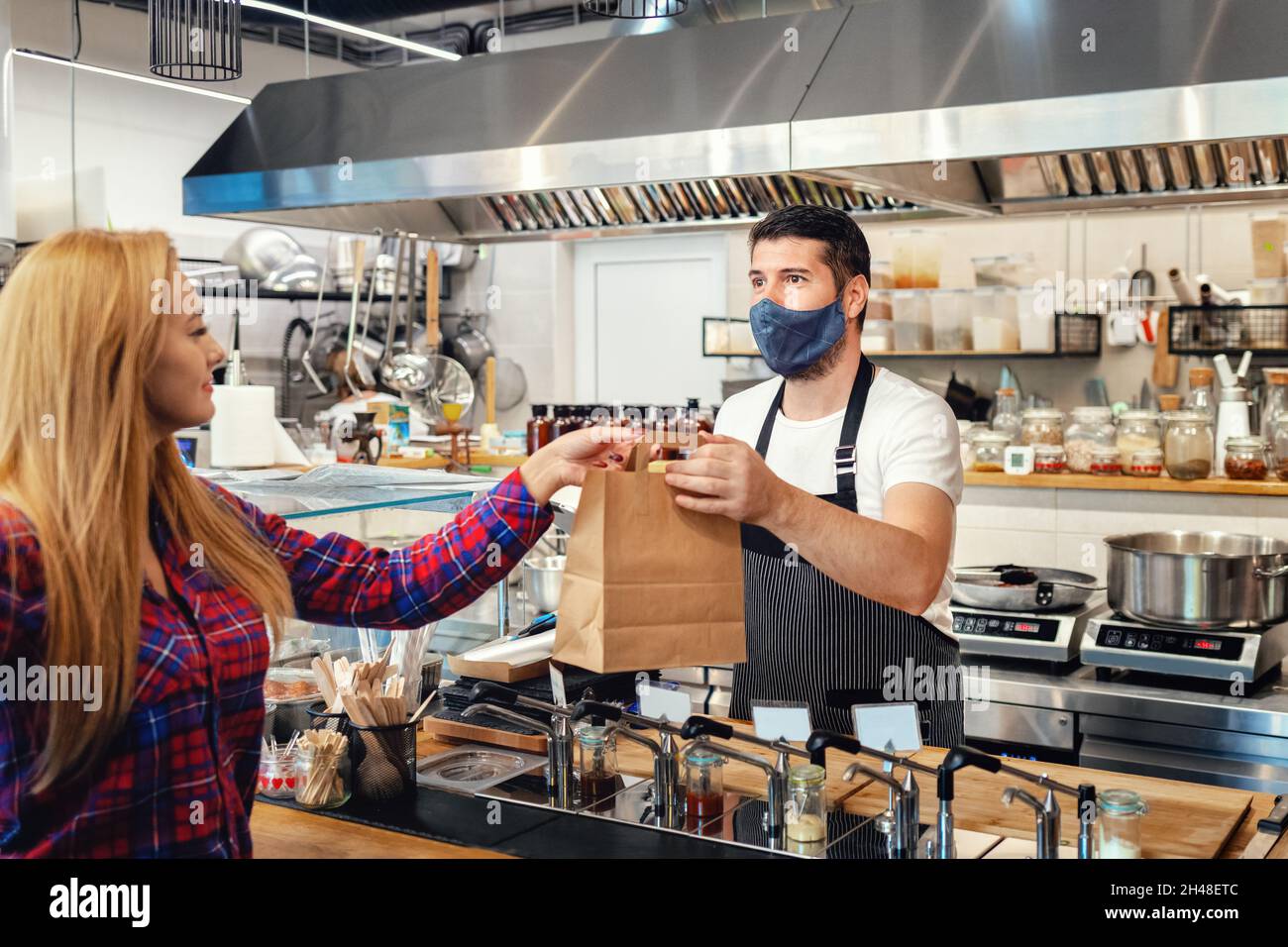 Proprietario con maschera facciale che serve cibo da asporto al cliente al banco in un piccolo ristorante per famiglie. Foto Stock