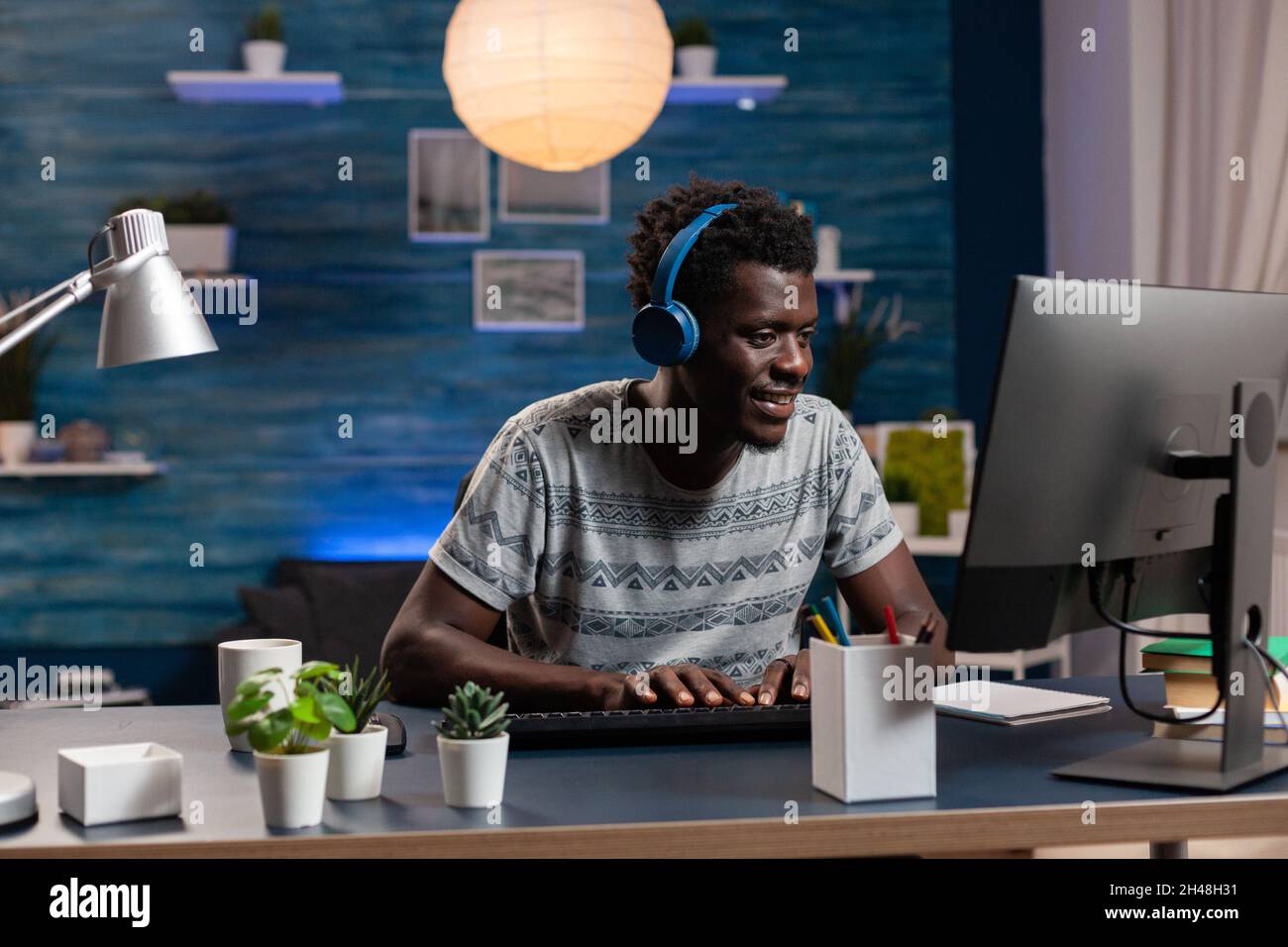 Dipendente afroamericano con cuffie digitando idee di marketing sul computer che lavora lontano da casa al progetto aziendale. Giovane uomo che naviga in linea le informazioni che analizzano il grafico finanziario Foto Stock