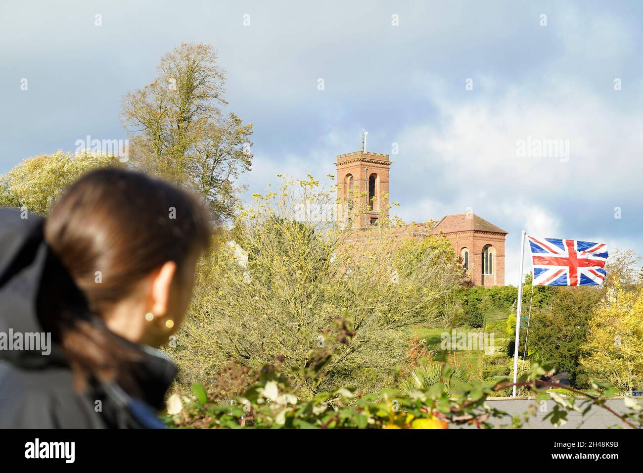 Vista posteriore di una donna che guarda una bandiera Union Jack che soffia nel vento con una chiesa britannica in lontananza. Foto Stock