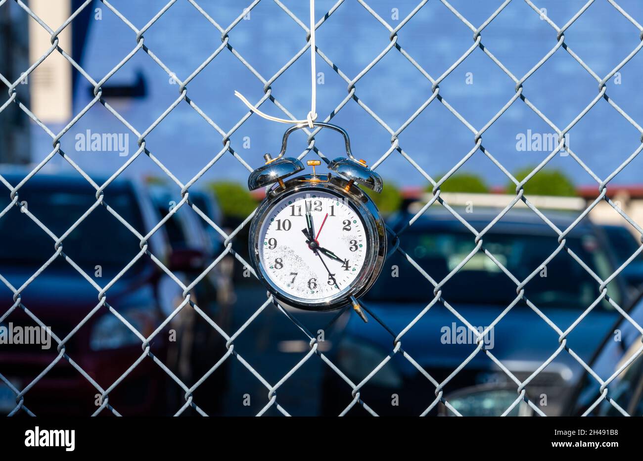Vecchio orologio sveglia appeso su rete metallica recinzione con le auto parcheggiate sul backgound Foto Stock